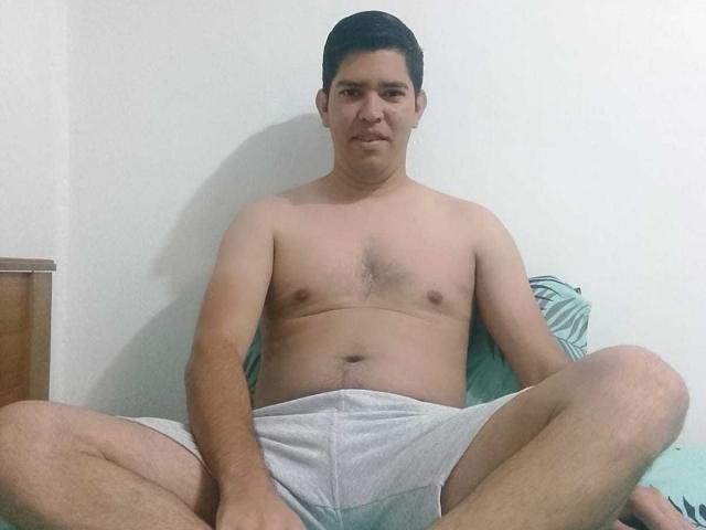 ChristopherRusso is sitting shirtless with a neutral expression, legs spread, wearing gray shorts, displaying a slightly chubby, hairy-chested, medium-sized ass, and dark hair.