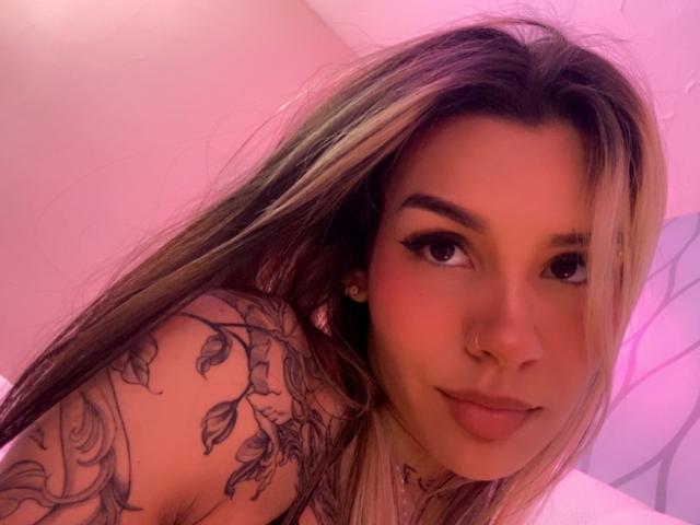 LenaBranch with blonde and brown hair, nose ring, and tattooed shoulder, smiling at the camera, showing her tits in a pink-lit room.