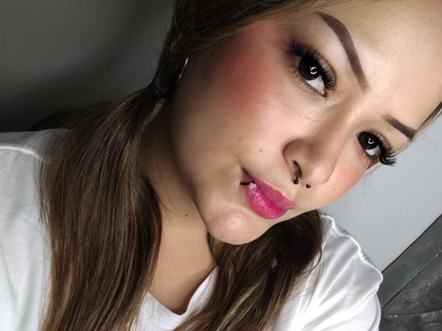 AllisonJustingb with puffy pink lips, brown eyes, and a nose ring, wearing a white shirt, has pink blush on her cheeks, and brown pigtails.