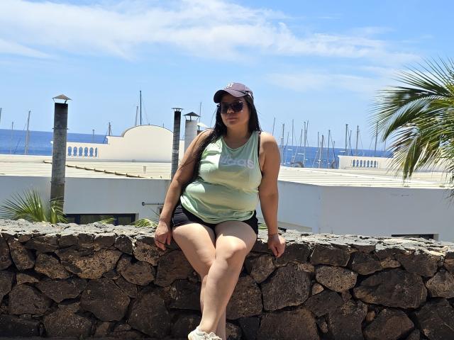 XBigTitxx with large tits, wearing a green tank top, black shorts, and a cap, sitting on a stone wall, wearing sunglasses, against a bright blue sky.