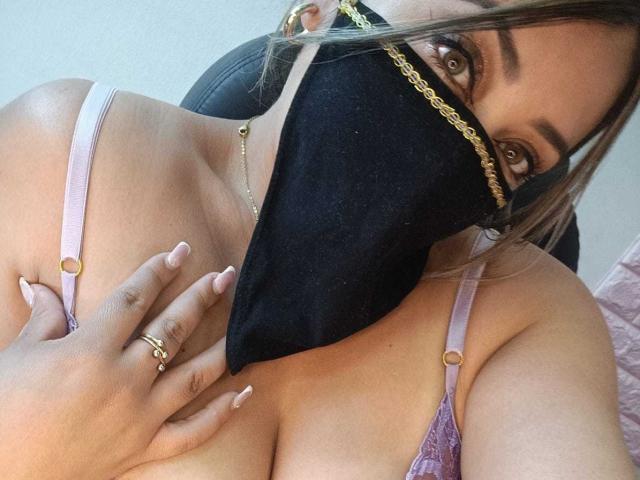 ZahraJalid with brown eyes, black mask, gold chain, pink bra, and rings, holding her tits, showing her pink-nail, tan-skinned, and blonde hair in a selfie.