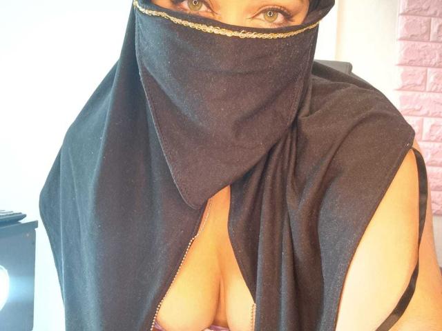 ZahraJalid with black mask covering her mouth and nose, black cloth exposing her cleavage, green eyes, and light skin, in a bright room, showing her bare shoulders and chest.