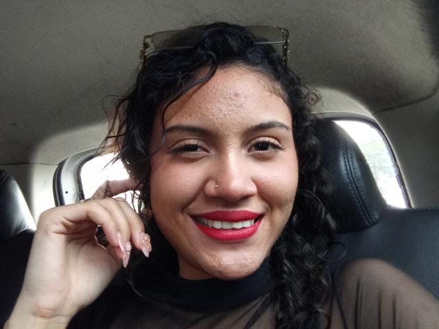 ValeryPoleth has curly black hair, medium skin, red lipstick, and a nose ring. She's smiling with red lips, showing teeth, and has a slightly pimply forehead. She's wearing a sheer top.