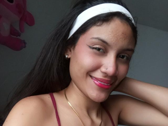 ValeryPoleth has a cute smile with pink lipstick, dark hair in a white headband, nose piercing, and eyeliner, wearing a red top and gold necklace, showing her small tits.