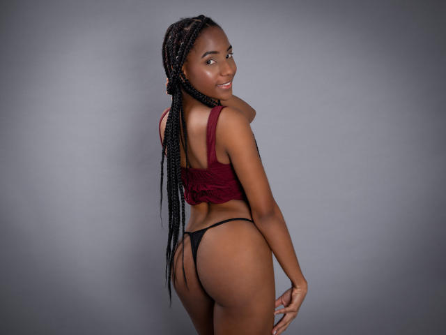 ChannelSweett with long black braids, red crop top, and black thong, showing her ass, smiling over her shoulder, standing against a gray background.