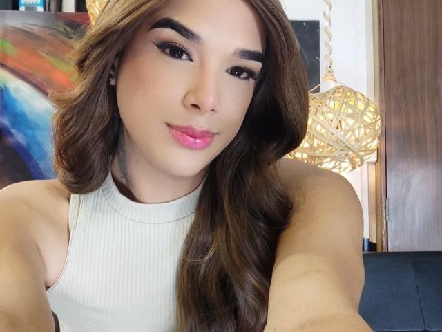 VictoriaAleia with long brown hair, pink lips, and a white sleeveless top, showing her smooth arms and a slight smile. Her tits are small, and she's looking at the camera.