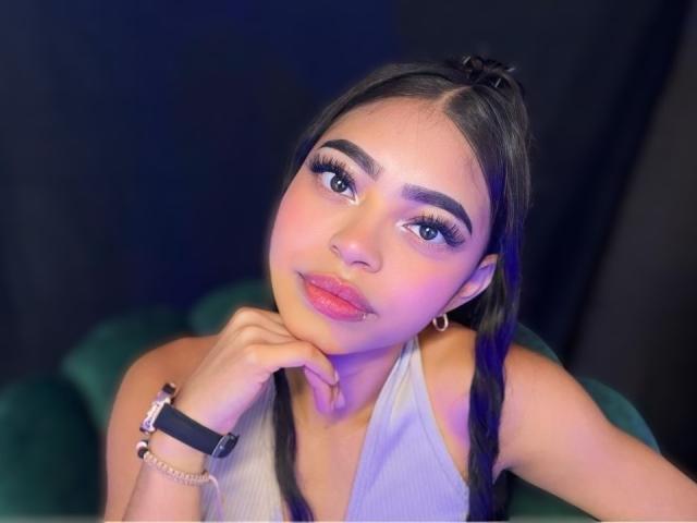 IsabellLaurent with dark hair in pigtails, wearing a white tank top, has her chin on her hand, looking up with pink lipstick, blue eyes, and a nose ring.