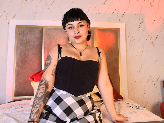 LilithVelvet met kort zwart haar, donkere lipstick, een zwarte top zonder bandjes, geruite broek, een getatoeëerde arm, zwarte oorbelletjes, een ketting, zittend op een bed, met rood en oranje licht..
