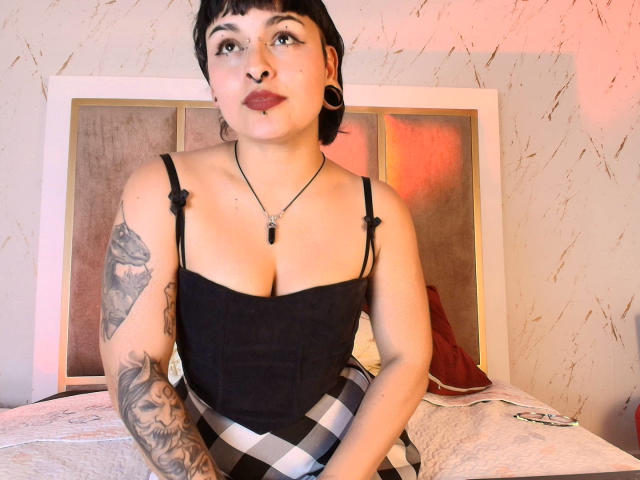 LilithVelvet, met kort zwart haar en tatoeages, draagt een zwarte corset en een geruite rok, waarbij haar borsten zichtbaar zijn, en heeft kleine zwarte oorbellen. Ze zit op een bed..
