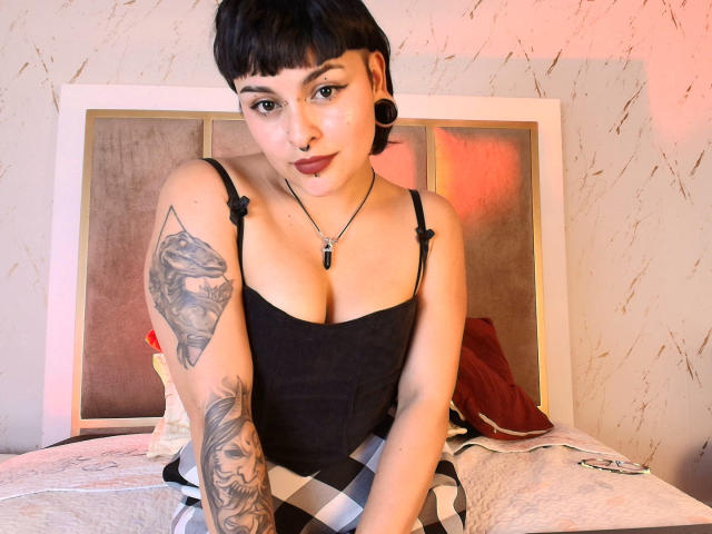 LilithVelvet, kurze schwarze Haare, schwarzes Tanktop, zeigt Brüste, tätowierter Arm, schwarze Ohrringe, Nasenring, Halskette, sitzt auf dem Bett, schaut in die Kamera, roter Lippenstift, karierter Rock..
