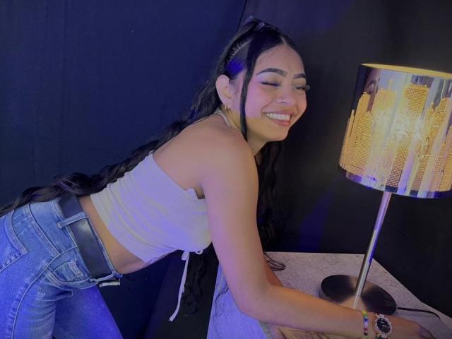IsabellLaurent with long black braids, smiling, wearing a white crop top and blue jeans, leaning on a table with a city-patterned lamp, showing her cute, smiling face.