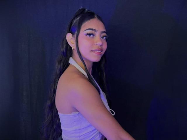 IsabellLaurent has long dark hair in a ponytail, wearing a white strapless top, showing her tits, with pink eyeshadow, smiling at the camera, against a dark background.
