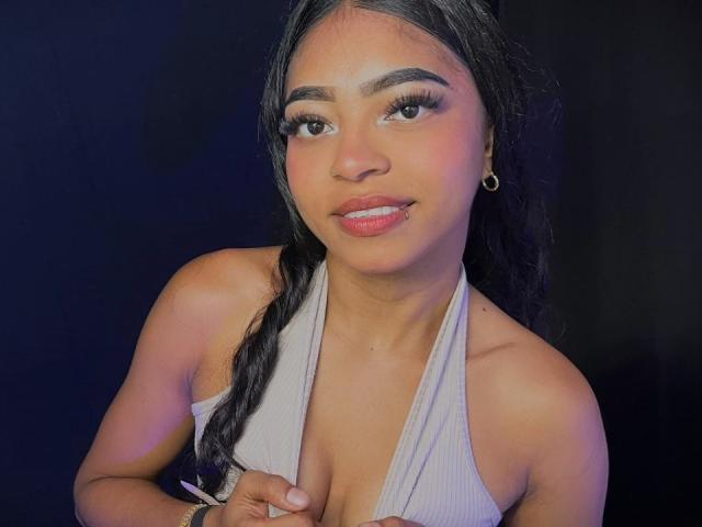 IsabellLaurent with long black hair in a braid, wearing a grey halter top, showing her small tits, and smiling with pink glossy lips, wearing gold hoop earrings.
