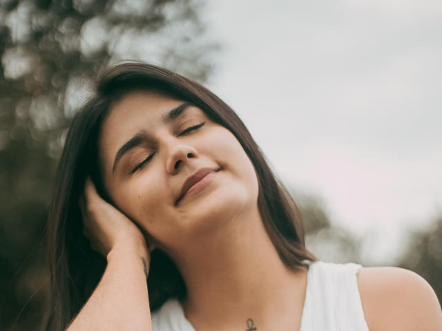 NathalyWalls with closed eyes, smiling slightly, holding her brown hair with one hand, wearing a white tank top, showing a small tattoo on her neck. Soft, natural light.