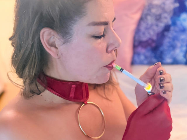 EmiliaMilf avec un collier et une bague rouges, suçant une sucette verte et blanche, ses seins exposés, cheveux bruns, piercing au nez et vernis à ongles noir, dans une chambre..
