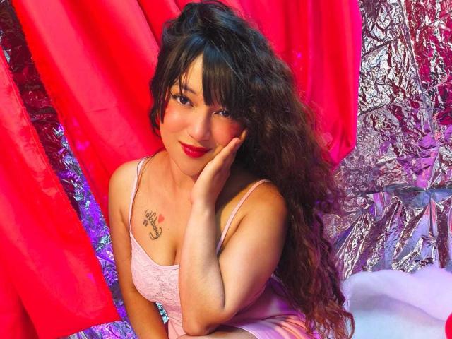 NicoleMartins with long curly brown hair, pink dress, red lipstick, hand on cheek, tattoo on left chest, looking at camera, sexy, pink and silver background.
