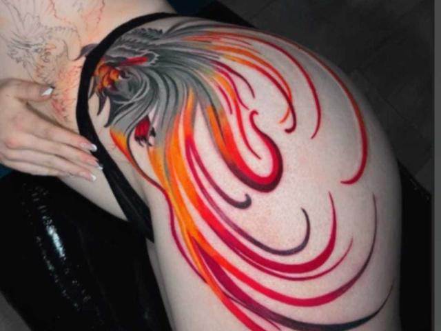 Tattoo on AliceCathyHot's shoulder. Vibrant, flowing design with a phoenix-like bird at the top. Bird's head has grey and red feathers, with an intense red eye. Bright orange, red, and pink swirling lines extend from the bird, covering the shoulder. AliceCathyHot's pale skin contrasts with the bold, colorful tattoo. Black strap of a bikini is visible on AliceCathyHot's right side. Tattoo artist uses thick, smooth lines and vivid colors, emphasizing motion and intensity.