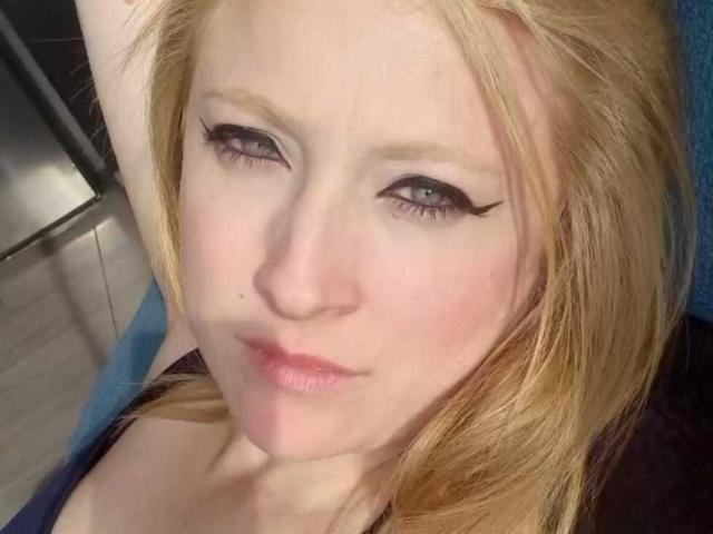 AliceCathyHot has pale skin, blonde hair, and winged black eyeliner. Her small tits are slightly visible under a dark top. She's looking up with a slight, confident smile.