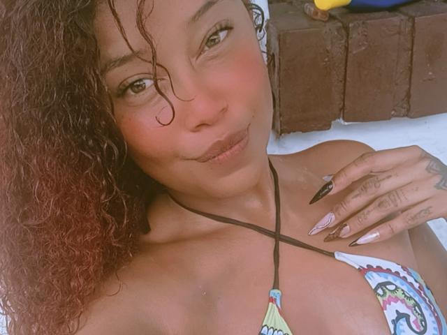 DalilaHeinz with curly brown hair, brown eyes, and a smile, wearing a colorful bikini, showing her tits and long, pointed nails with tattoos on her hand.