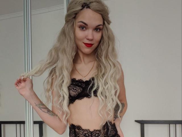 FloraAmber mit langem, blonden, gewellten Haar, einem schwarzen Spitzen-BH und passender Unterwäsche, die ihre Haare hält, rotem Lippenstift, Tattoos am rechten Arm, lächelnd, stehend in einem Raum..