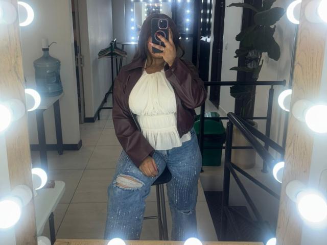 SofyaZaens sits in a mirror-lined room, taking a selfie with her phone. She's wearing a dark brown leather jacket over a white, ruffled blouse, and ripped blue jeans. Her left knee is bent, foot resting on a stool. Bright, round mirror lights frame the mirror. Her dark hair is loose, partially covering her face. The background shows a water dispenser and green bin. The room is well-lit, highlighting her casual, stylish look.