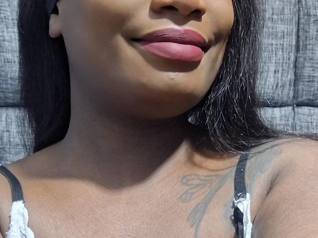 AlissaJordan's face with full pink lips, brown skin, and long black hair. She's wearing a white lace top, showing a tattoo on her right shoulder.
