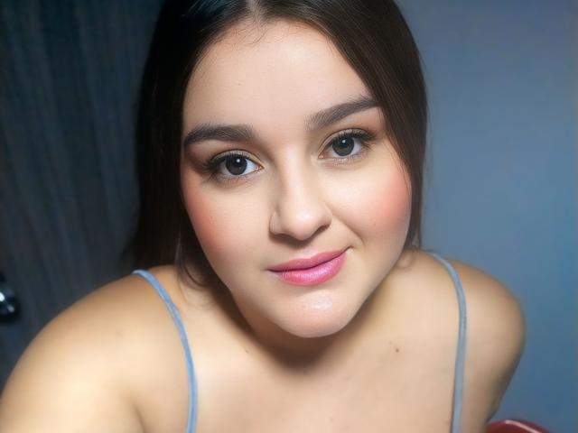 TessieMira with pale skin, brown eyes, pink lips, and straight brown hair, wearing a blue spaghetti-strap top, showing a subtle smile. Boobs visible in the upper frame.