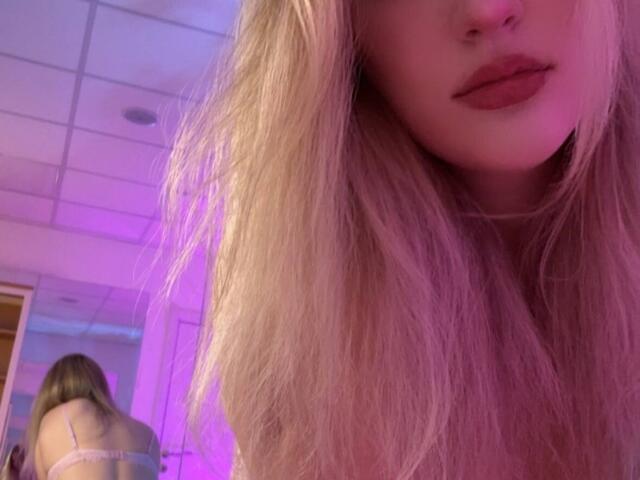 Close-up of Synrix with long blonde hair, pinkish-purple light illuminating her. Synrix's face partially visible, lips painted in dark red. Reflection in background shows Synrix's back, wearing a pink bra, standing in a room with white ceiling tiles. Soft, blurred texture to hair, contrasting with sharp reflection. Synrix's skin is fair, hair slightly messy. Image focuses on her hair and lips, with the reflection providing context. Slightly tilted perspective.