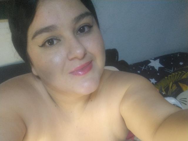 TessieMira with shiny pink lips, black eyeliner, and short black hair, showing bare shoulders, slightly smiling, and looking at the camera. Her tits are not visible.