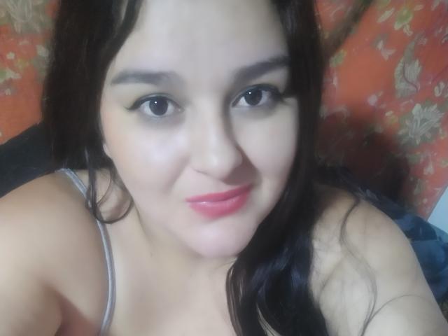 TessieMira with dark hair, pale skin, black eyeliner, red lipstick, and a slight smile, showing her tits in a gray top, looking at the camera.