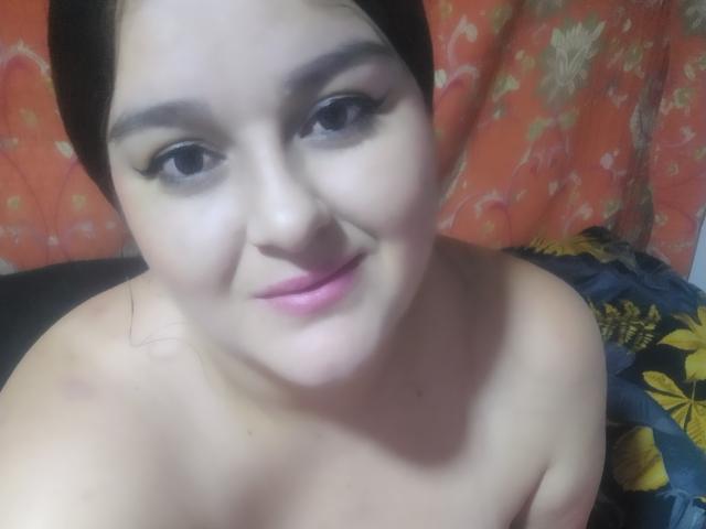 TessieMira with dark hair, heavy eyeliner, pink lipstick, and a slight smile, topless, showing her bare shoulders and upper chest, with a soft, pale skin tone.