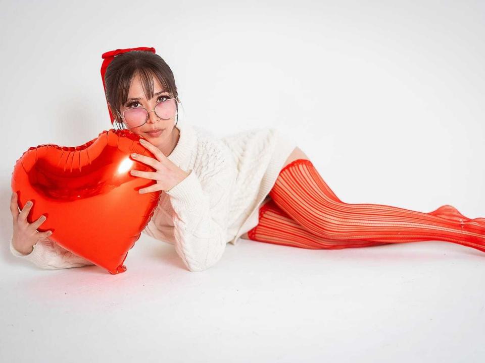 Luuna69 with red-striped knee socks, white sweater, and glasses, holding a shiny red heart, lying on white floor, looking at camera.