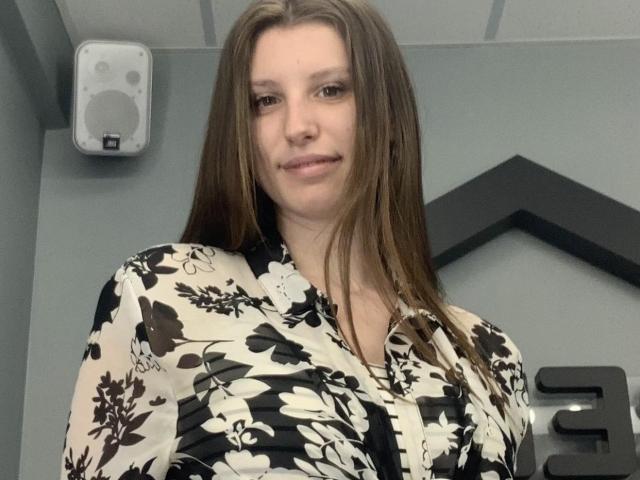 LilianaShine with long brown hair, wearing a black and white floral shirt, has a slight smile, and her tits are partially visible, showing some cleavage.