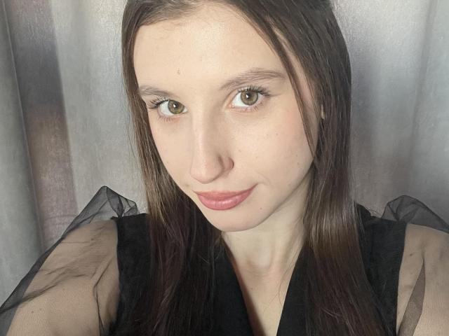 LilianaShine has brown eyes, light skin, and long brown hair. She's wearing a black sheer top with pink lipstick. Her small tits are partially visible. She's smiling.