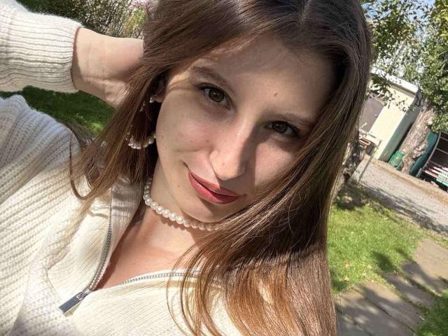 LilianaShine with brown hair, pearl necklace, white zip-up sweater, and red lipstick, smiling at the camera, hand in hair, sunlight highlighting her face.