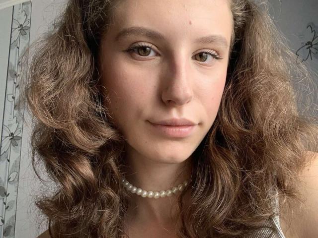 LilianaShine has wavy brown hair, green eyes, and a pearl necklace. Her slightly smiling face shows a cute, natural look. Boobs not visible, focus on her face.