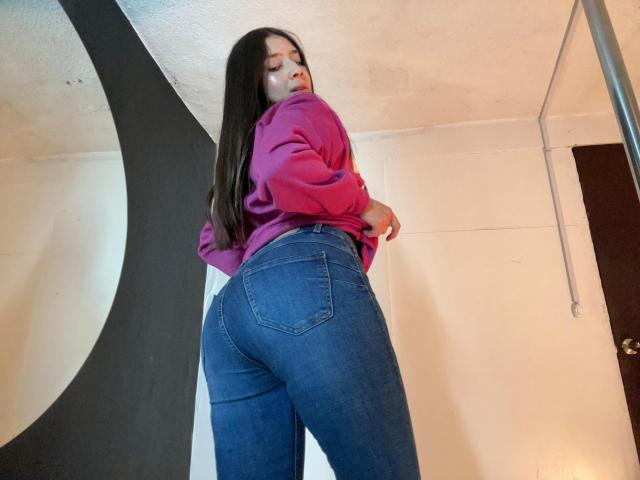 EmmaLombardi with long brown hair, wearing a pink sweatshirt and tight blue jeans, showing her big ass and looking back with a slight smile. Low angle, indoors.