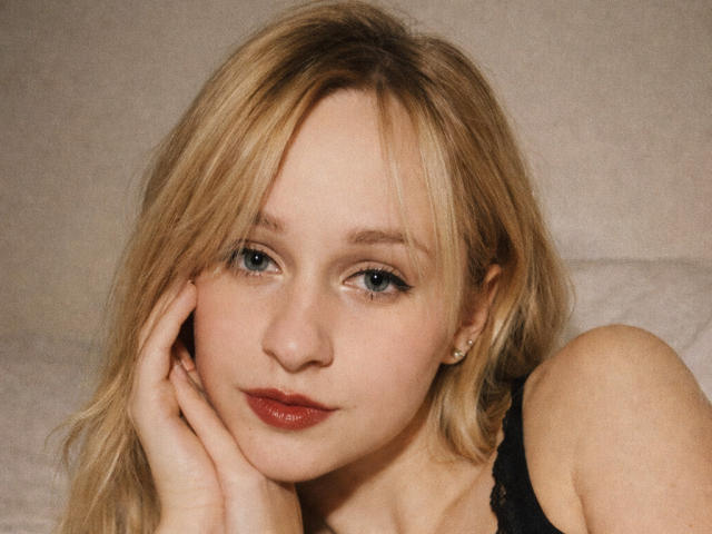 FlyFreedom has blonde hair, blue eyes, and a fair skin tone, with a slight pout and red lipstick. She's touching her cheek, wearing black lace, and looking serious.