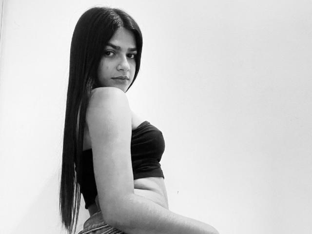 DeviColens with long dark hair, black strapless top, and visible midriff, looking over her shoulder with a slight smile, in a black-and-white photo. Her ass is slightly visible.