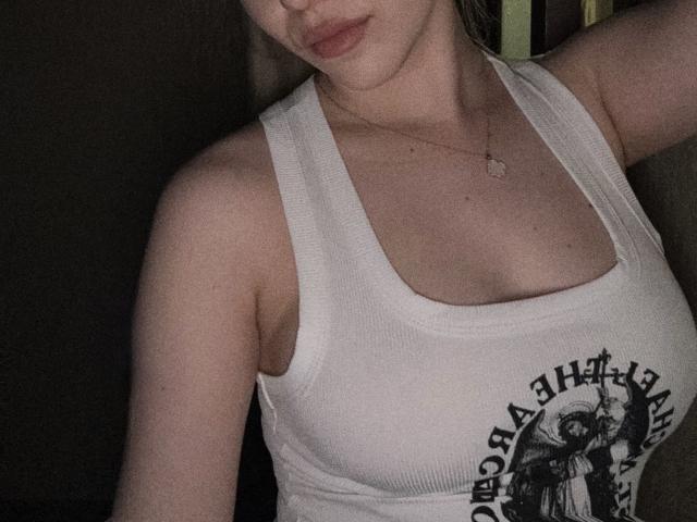 SugarrX trägt ein weißes, geripptes Tanktop mit einem Aufdruck, der ihre kleinen Brüste und ihre helle Haut zeigt. Sie trägt eine zarte Halskette und hat den Kopf leicht geneigt..