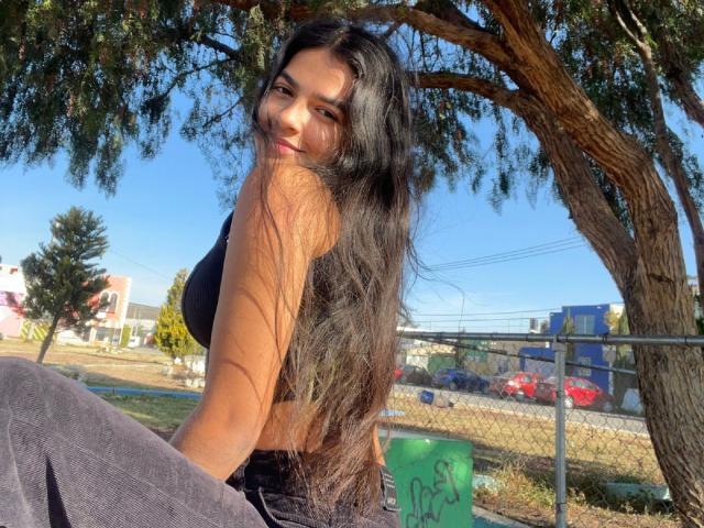 DeviColens with long black hair, smiling, wearing a black sports bra and dark pants, sitting under a tree, blue sky, and a chain-link fence in the background.