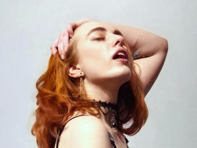 RubyLust-hot with red hair, eyes closed, mouth open, hand in hair, black choker, earrings, and a nose piercing, looking sexy and slightly aroused.