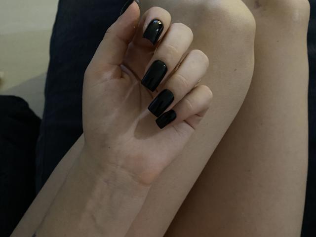 Olieri's hand with black, glossy nail polish is gripping her knee. Her pale skin is visible. The image focuses on her fingers and knee.