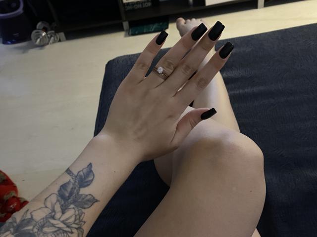 Olieri's hand with black nails and a diamond ring, resting on her bare legs, showing a floral tattoo on her wrist. Her feet are crossed on a dark blue surface.