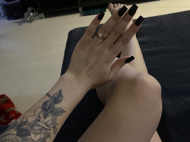 Olieri's hands with black nails, ring, and tattoo, resting on her bare legs, showing a diamond ring. Her ass is partially visible.