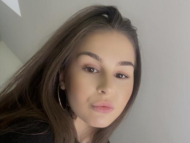 AriaDevone has light skin, brown eyes, and brown hair with a slight ponytail. She's wearing thin hoop earrings and has pinkish lips, looking slightly to the left.