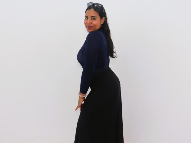 LiliLopez has dark blue top, black skirt, and long black hair, looking back with a smile, wearing sunglasses on her head, standing against a white wall.