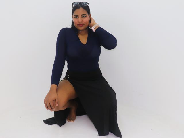 LiliLopez with dark skin, wearing a navy long-sleeve top and black skirt with a high slit, squatting, black socks, glasses on head, confident smile, hand in hair.