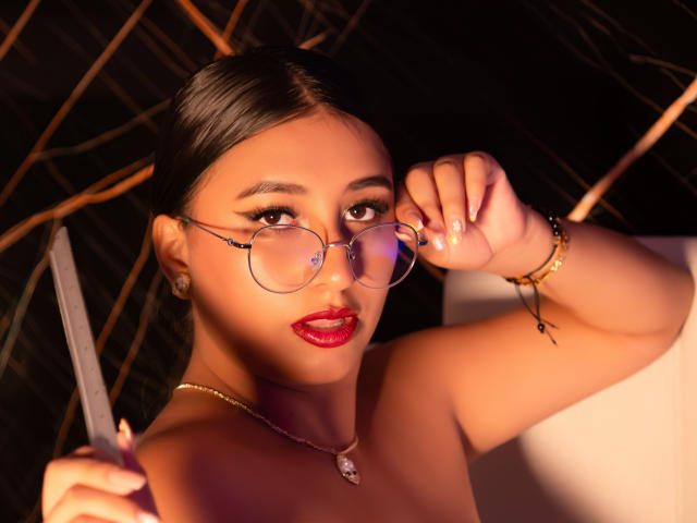 Catalina Rivero, com cabelo escuro, batom vermelho e óculos, segurando uma faca na mão direita, usando um colar e um bracelete, tem um visual sexy e sedutor..