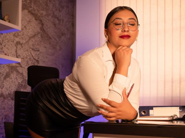 Catalina Rivero, avec des lunettes, une chemise blanche et une jupe en cuir noir, appuyée sur un bureau, la main sur le menton, tenant un crayon, rouge à lèvres, cheveux courts et foncés, confiante, séduisante, avec un léger sourire..